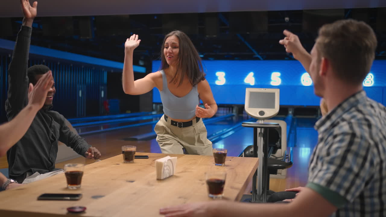 Una donna bruna felice in una sala da bowling corre gioiosamente dalle sue amiche per celebrare lo strike. Condividi la vittoria con gli amici. Gruppo di amici multietnico