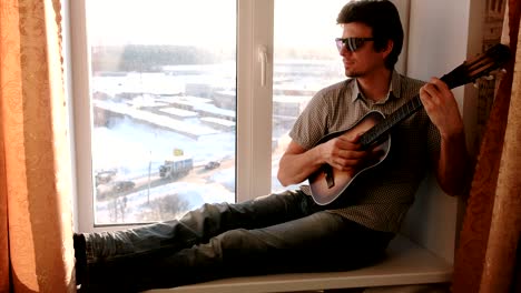 playing a musical instrument. man in sunglasses is playing the guitar and singing sitting in windowsill.