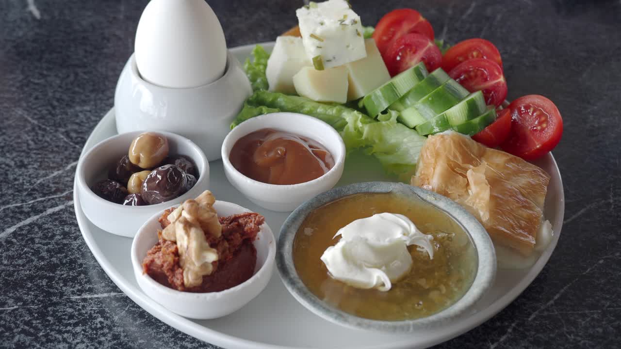 A Turkish breakfast plate with eggs, cheese, olives, tomatoes, cucumber, honey, butter, jam, walnuts, pastry, and bread