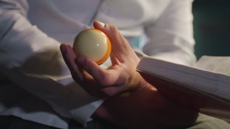 close-up shot of person in white shirt, face not visible, holding an open book and billiard ball in one hand. focused gesture, dim lighting, cinematic mood, and contemplative atmosphere
