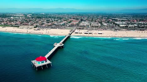 Vista-De-Drones-Volando-Alrededor-Del-Final-Del-Muelle-En-Huntington-Beach,-California,-Con-Una-Excelente-Vista-De-Las-Montañas,-La-Playa,-Las-Olas,-El-Surf-E-Incluso-Algunas-Personas-Caminando