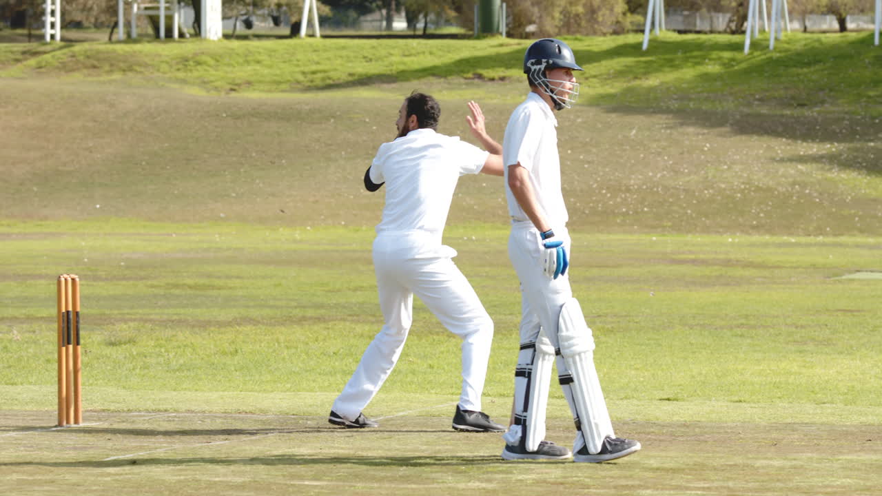 Two multiracial male cricket players playing cricket, bowler throwing the ball on pitch