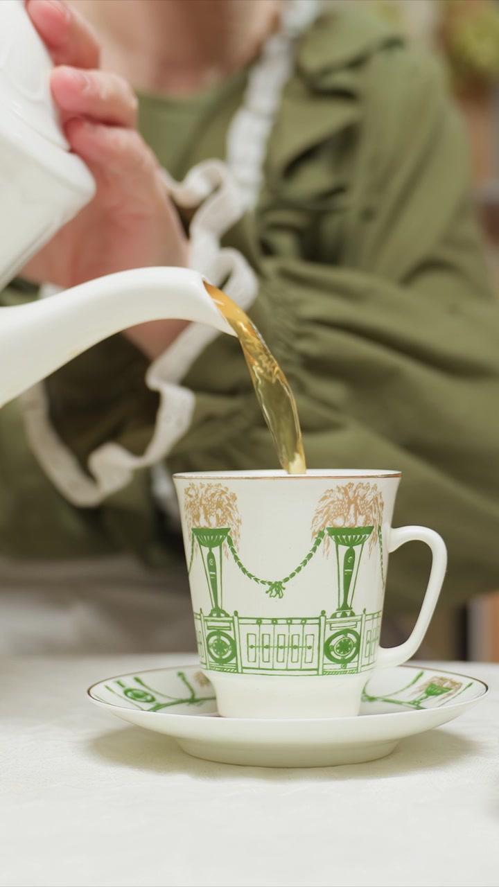 Una donna in abito verde versa il tè in una tazza da tè e piattino dal design intricato. La tazza presenta motivi architettonici con colonne, catene, vasi. Un oggetto rosa poggia lì vicino su un altro piatto.