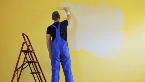 painter doing the makeover indoors. male worker painting wall with a paint roller next to the ladder. improvement of a room interior. back view.