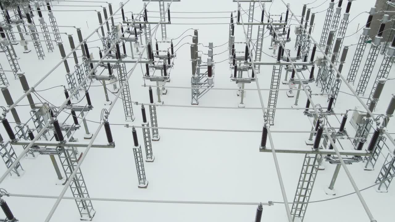 Aerial backward flight over a frozen electrical substation reveals snow covered insulators, cables, and grid structures resting on untouched white ground during a cold winter day