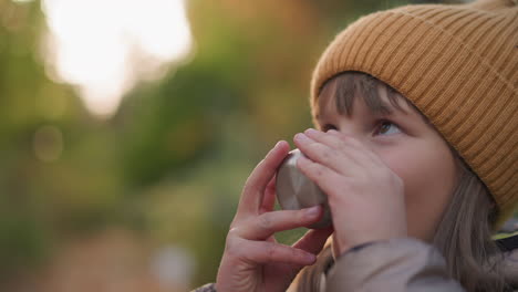 close up of adult hand offering cup of juice to child pressing lips to rim and drinking eagerly, set against warm autumn bokeh background, capturing nurturing moment of care and refreshment