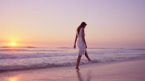 Hermosa-Mujer-Con-Vestido-Blanco-Caminando-A-Lo-Largo-De-La-Costa-En-La-Playa-Al-Atardecer-En-Cámara-Lenta-Dragón-Rojo