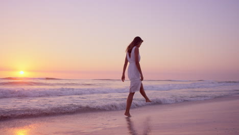 beautiful woman wearing white dress walking along shore line on  beach at sunset in slow motion red dragon