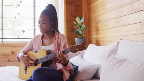 Feliz-Mujer-Afroamericana-Sentada-En-El-Sofá-Y-Tocando-La-Guitarra-En-Una-Cabaña-De-Madera,-Cámara-Lenta