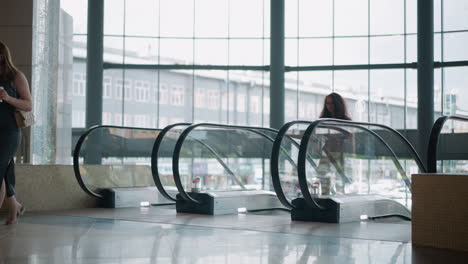 mujer saliendo de la escalera mecánica en un edificio moderno con paredes de vidrio con vista al paisaje urbano al aire libre, coches estacionados y otros edificios en el fondo, la escena captura la arquitectura urbana, el contraste interior-exterior