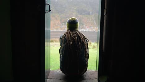 A-man-with-dreadlocks-and-wearing-hiking-gear-sits-on-a-step-in-a-narrow-doorway-of-a-bothy-in-the-Highlands-of-Scotland-to-enjoy-the-view-outdoors