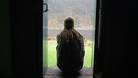 un hombre con rastas y equipo de senderismo se sienta en un escalón en una puerta estrecha de un bothy en las tierras altas de escocia para disfrutar de la vista al aire libre