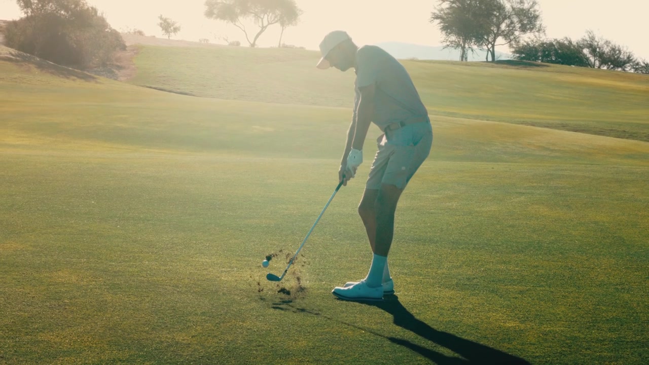 A lone golfer swings wedge and hits behind ball with big divot into bright sun flare, backlit against the desert at golden hour in slow motion, a dramatic sequence for sports and lifestyle edits.