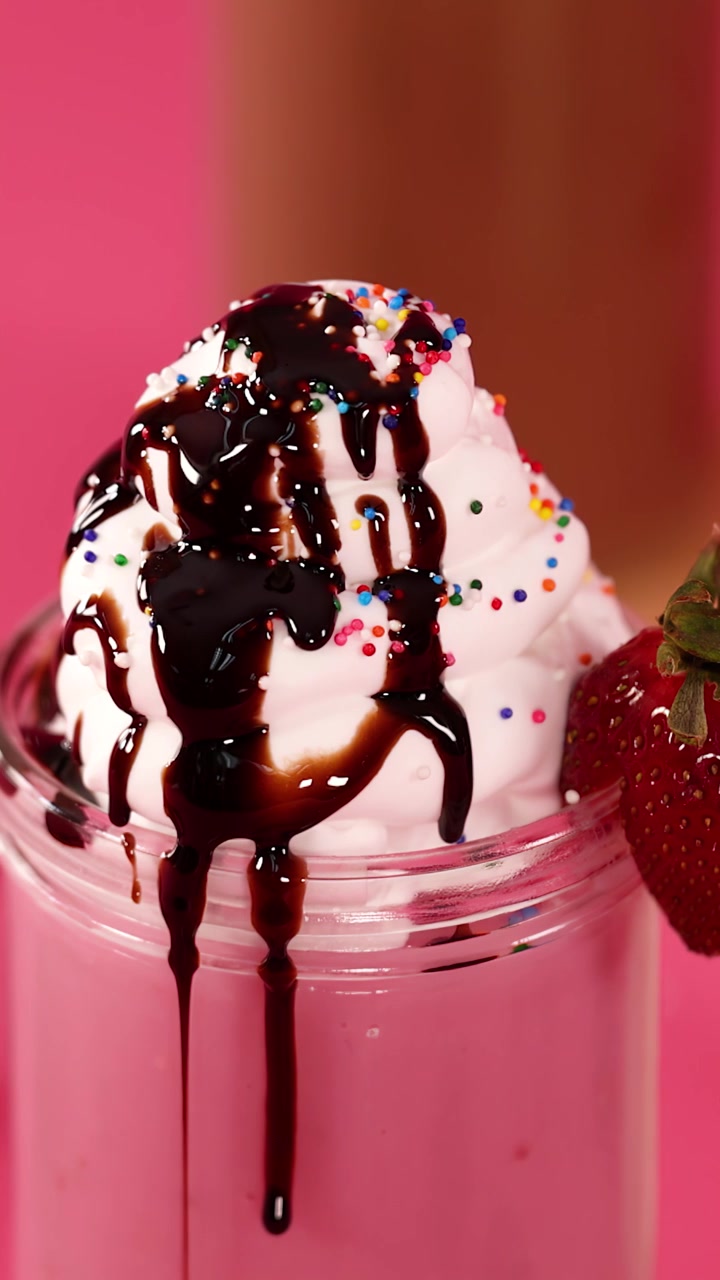 Frullato rosa alla fragola guarnito con panna montata, sciroppo al cioccolato, codette colorate e una fragola, sotto una luce brillante.