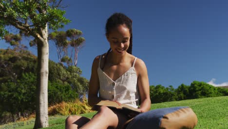 African american woman sitting on grass reading book in park