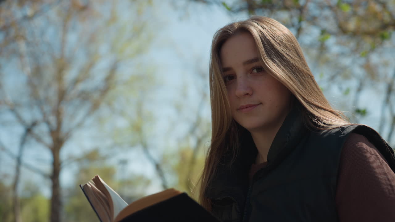 Mujer absorta en un libro en el exterior, mujer caucásica leyendo tranquilamente en un entorno primaveral, joven caucásica relajándose y reflexionando mientras lee bajo la luz del sol primaveral en un parque.
