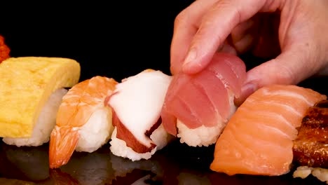 a hand delicately picks up various pieces of nigiri sushi from a neatly arranged platter under bright, even lighting with a glossy black background