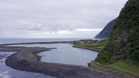 vista de drones que revela un pueblo costero, exuberante paisaje de acantilados verdes, laguna, fajã de santo cristo, isla de são jorge, las azores, portugal