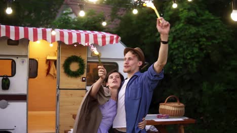 Young-Couple-Embracing-Standing-In-Front-Trailer-They-Living-In-With-Sparklers-Celebrating,-Waving-Hands-With-Fireworks-Sticks