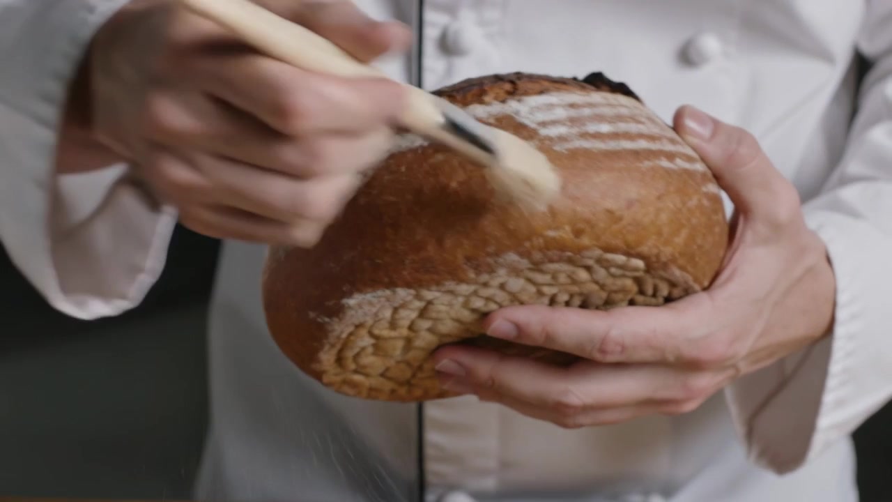 Presenting loaf, chef in white chef jacket brushing loaf with pastry brush, dusting flour in studio