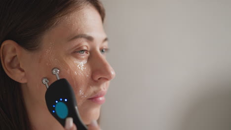 female does microcurrent procedure for skin lifting. brunette lady makes massage movements on face skin near eyes using microcurrent in bathroom