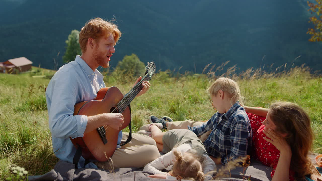 Father playing guitar nature. Happy family singing sitting blanket at picnic.