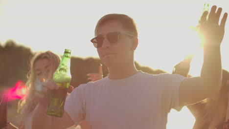 joven en camiseta blanca y gafas de sol de moda negras está bailando con sus mejores amigos en la fiesta al aire libre con cerveza. disfruta de esta noche de verano al atardecer.