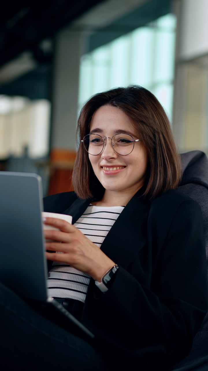 Caucasian girl sits comfortably in bean bag chair holding a laptop. Relaxed woman works on laptop and drinks coffee. Vertical video.