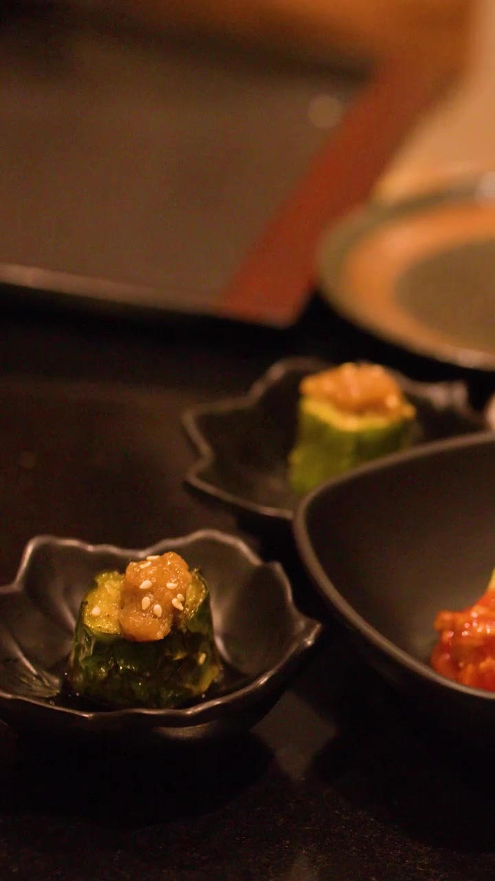 Close-up de mãos arrumando pequenos pratos de quiabo e kimchi em uma mesa escura de restaurante
