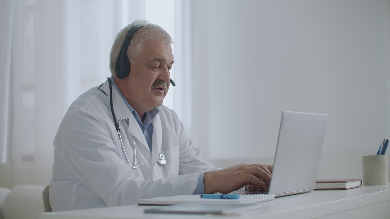 el médico está hablando con el paciente en línea por videollamada en el portátil escuchando por auriculares y llenando el informe médico trabajando de forma remota