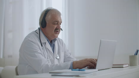 male doctor is talking with patient online by video call on laptop listening by headphones and filling medical report working remotely