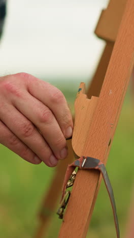 una imagen en primer plano de la mano de un hombre ajustando delicadamente un trípode de madera al aire libre en un campo cubierto de hierba. en el fondo, la pierna de una mujer es visible, añadiendo una atmósfera serena y enfocada a la escena