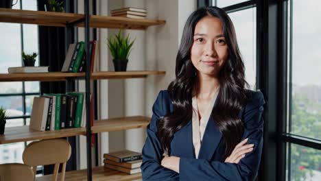 young asian businesswoman in elegant suit stands confidently with arms crossed in a contemporary office, exuding professionalism and success
