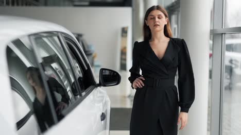 hermosa joven con un vestido sexy se acerca al auto nuevo y mira eróticamente al salón