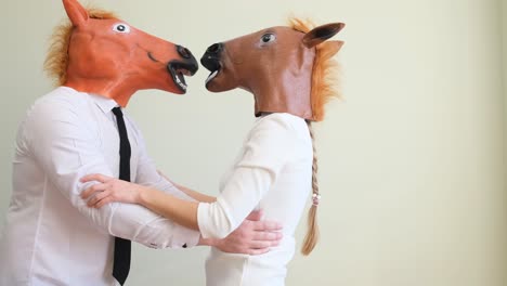 A Man and a Woman in White Shirts and Horse Masks Stand on a White and Kiss. A man and a woman in white shirts and horse masks stand on a white and kiss. Amazing fun. Humor concept