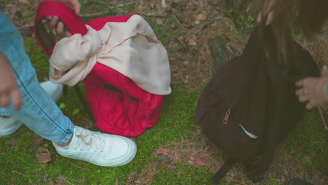 close-up of ladies in jeans and sneakers retrieving backpacks from the forest floor, carefully zipping them up before continuing their hike, surrounded by pine needles and green moss