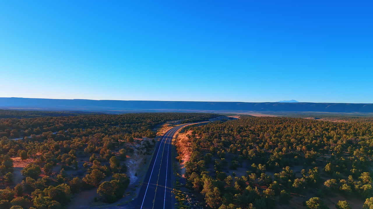 Desert highway approaching canyon rim. A long desert road leads toward the canyon rim. The winding path cuts through forested terrain toward distant cliffs