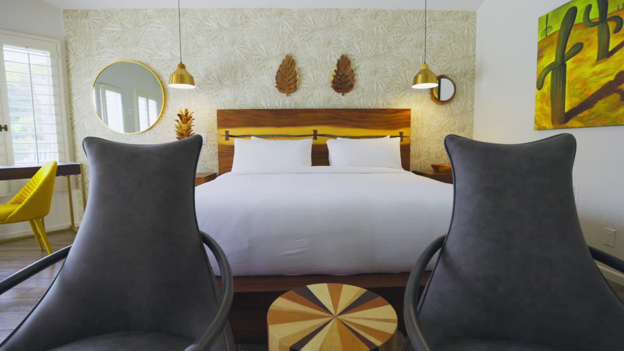 Two chairs in a hotel room with a large bed with white sheets, tracking wide shot
