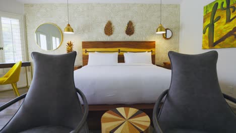 two chairs in a hotel room with a large bed with white sheets, tracking wide shot