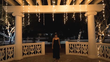 young brunette girl in a black dress advancing towards the camera in a nice stage with garlands of warm lights