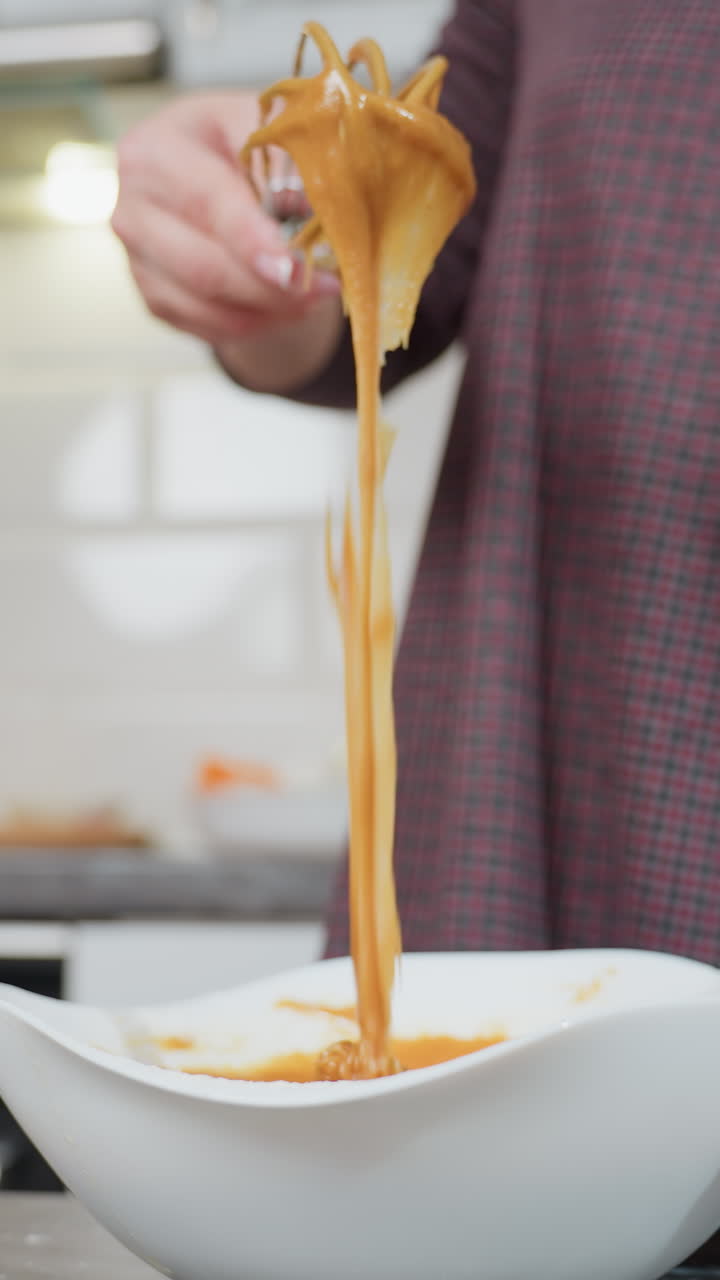 Close-up of woman in plaid dress lifting thick caramel-colored batter with whisk as it slowly drips back into white bowl, kitchen background with soft lighting, baking tools, and blurred cookbook