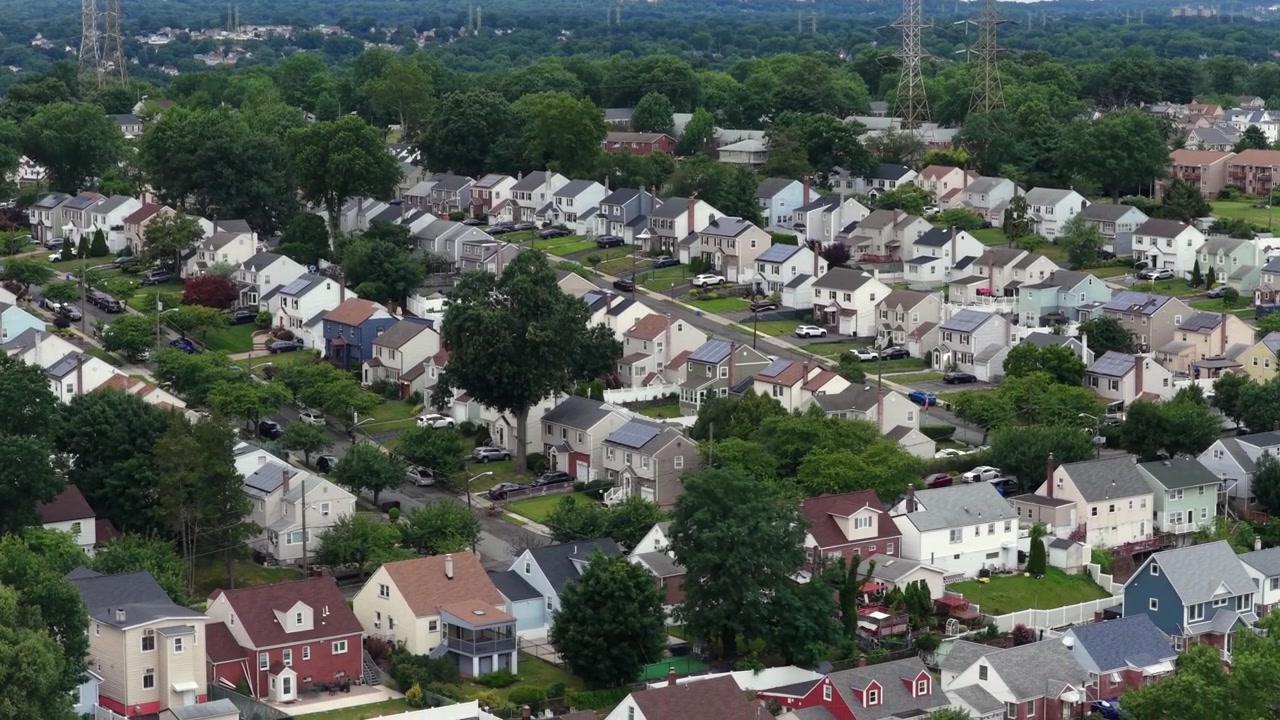 Case americane colorate a due piani con pannelli solari sul tetto fiancheggiano una griglia di tranquille strade suburbane, una fitta chioma di alberi verdi ombreggia vialetti e molte auto parcheggiate sotto. Ripresa aerea grandangolare con drone di un sobborgo americano.