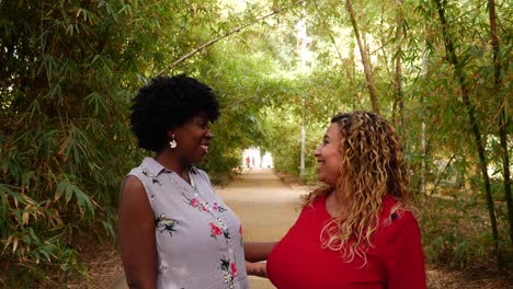 slow-motion medium shot of two women sharing a laugh and a shoulder hug while on a trail in a bamboo arboretum, evoking themes of friendship, diversity, womanhood, nature, warmth, and togetherness