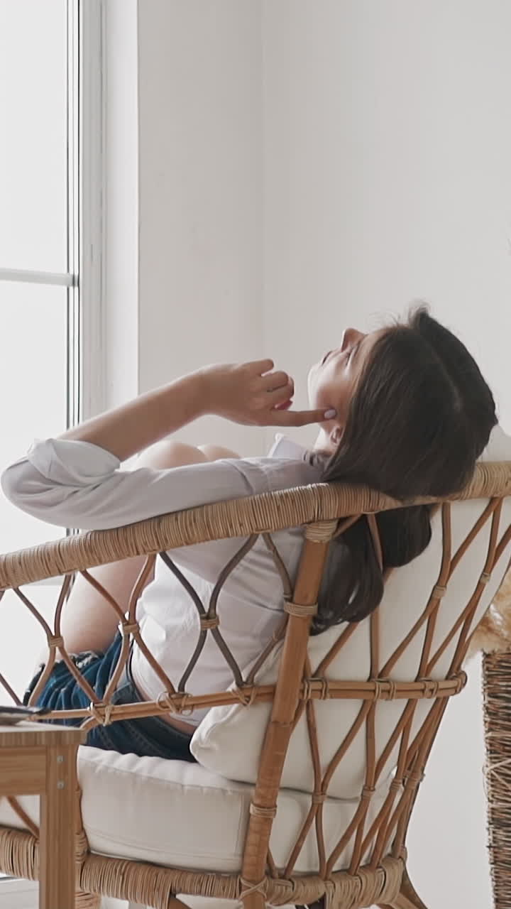 Lazy young woman artist in white shirt waits for inspiration sitting in rattan armchair near easel with picture in studio backside view slow motion