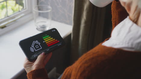 senior woman using app to measure energy efficiency standing by radiator at home