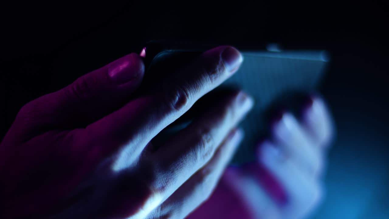 Thumb scrolling on a smartphone in landscape close up in a dark room with purple and blue lighting soft focus