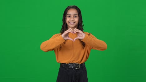 happy beautiful african american teen girl showing heart gesture looking at camera smiling. portrait of confident positive adolescent teenager posing on green screen chromakey. love concept.