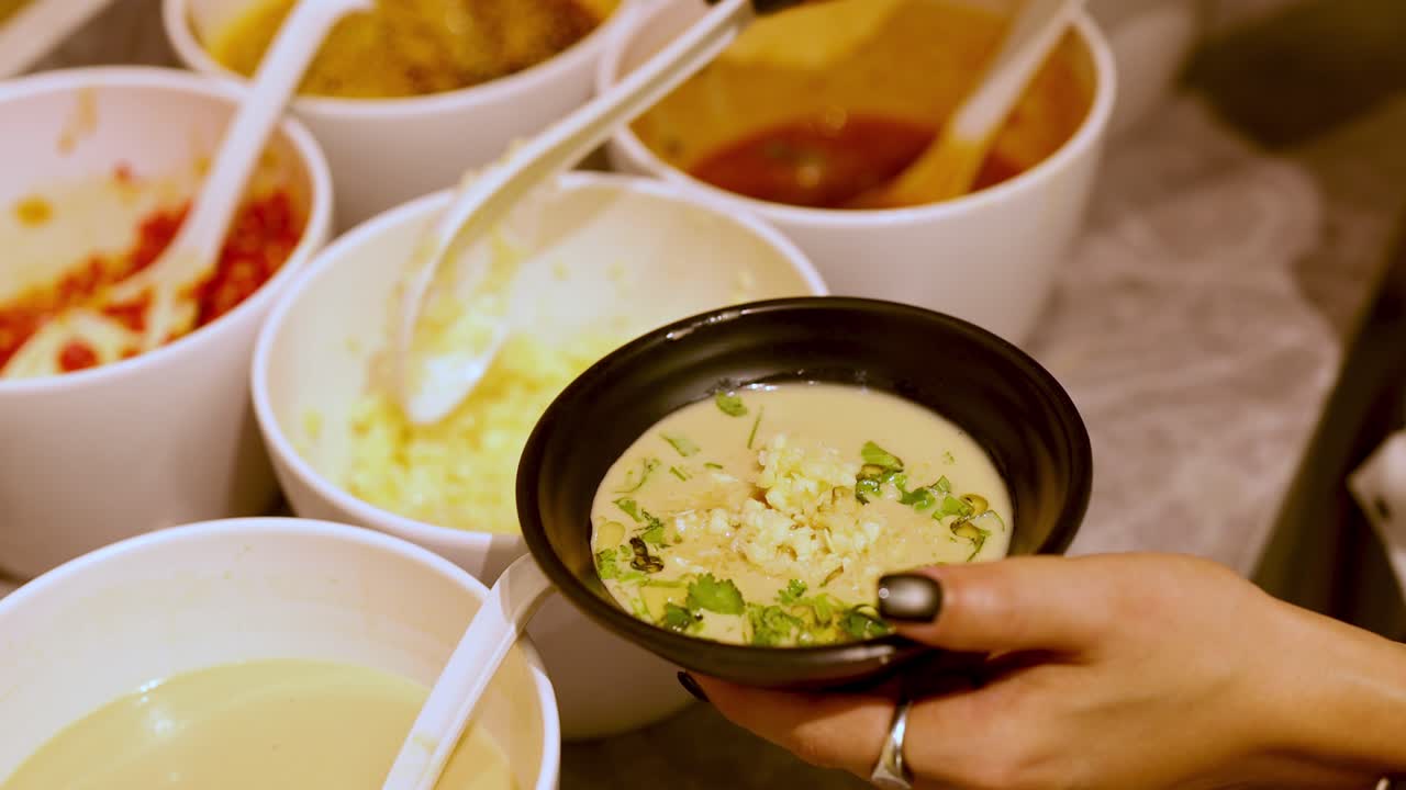 Hand scoops condiments into bowl at hotpot sauce bar under warm, indoor lighting