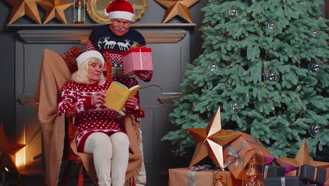 Senior-grandmother-presenting-Christmas-gift-box-to-surprised-grandfather-while-she-reading-book