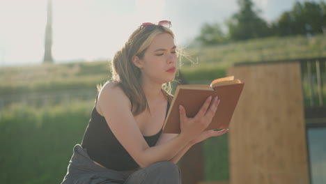 woman seated , reading a book as the wind blows her hair and pages, her hand rests on the book with a black bag nearby, and the background features a blurred grassy hill in the sunlight